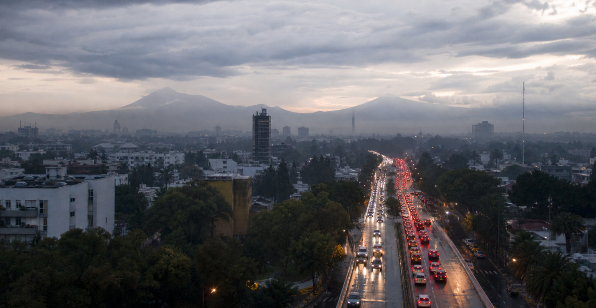 Tráfico vehicular en una avenida de la Ciudad de México al amanecer, con pavimento mojado y cielo nublado.