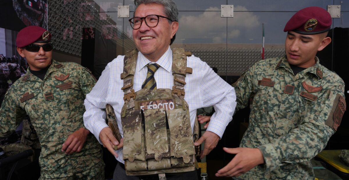 Ricardo Monreal prueba un chaleco táctico acompañado de dos militares durante una exhibición.